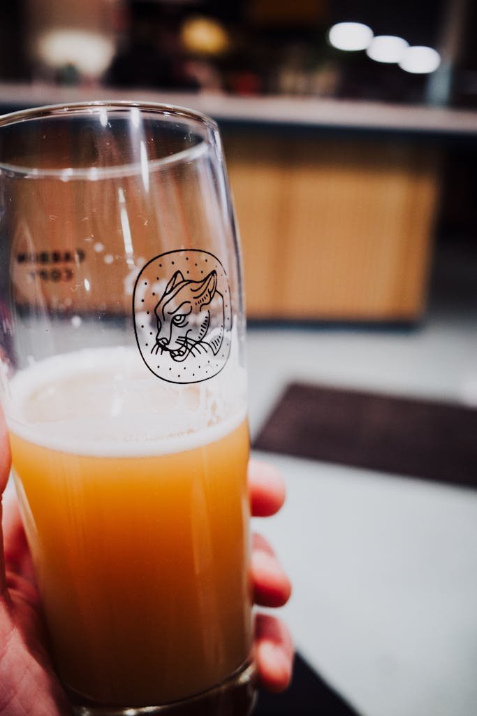 Close-up image of a hand holding a craft beer glass with a unique logo in a cozy bar setting.